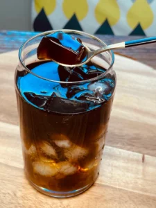 Glass of black tea with lychee jelly cubes over ice, showing rich amber gradient and blue reflections on wooden surface.